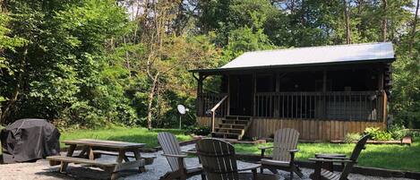 A rustic cabin with a covered porch, picnic tables, and Adirondack chairs in a wooded area.