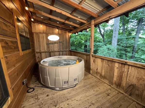 A wooden cabin with a hot tub, a wooden deck, and a view of a forest.