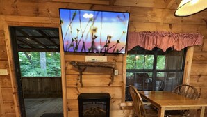 Interior de una cabaña de madera con chimenea, televisor montado en la pared, mesa de comedor y sillas.