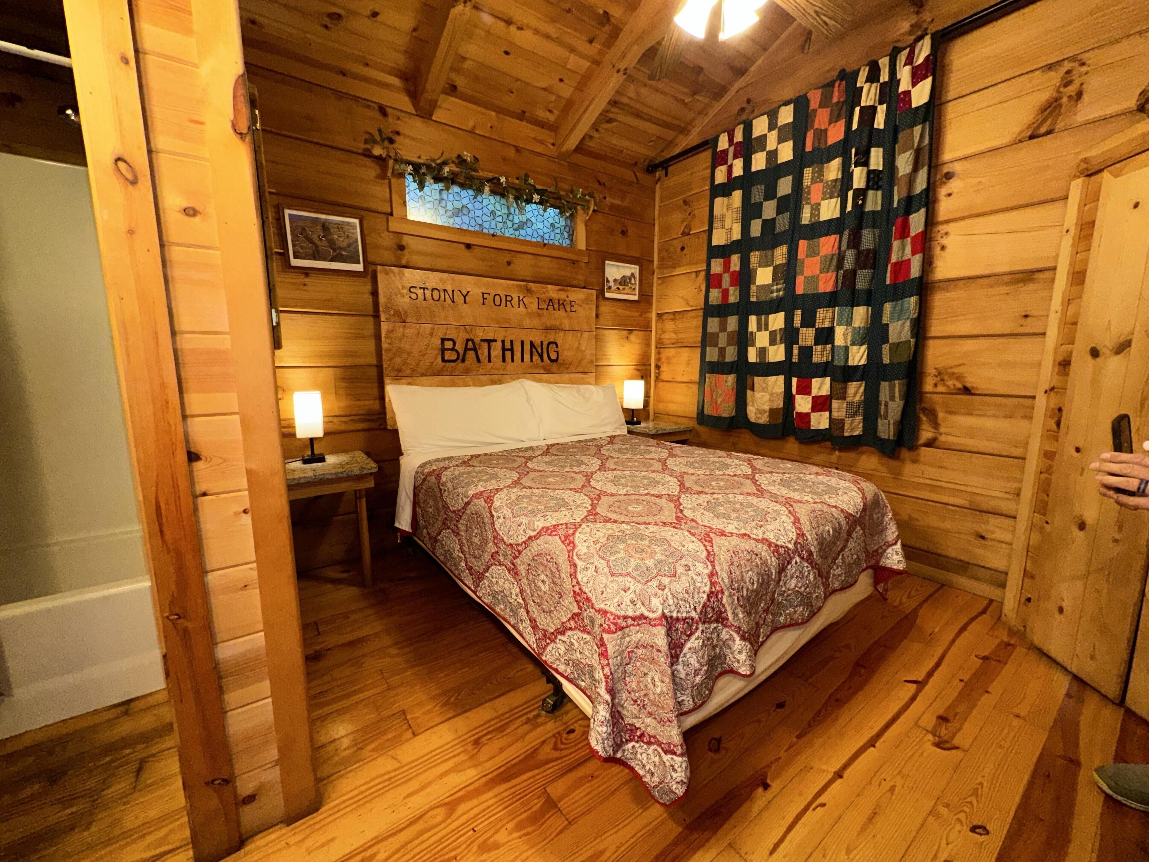 A wooden cabin room with a bed, a quilt, a sign reading 'Stony Fork Lake BATHING', and a quilted curtain.