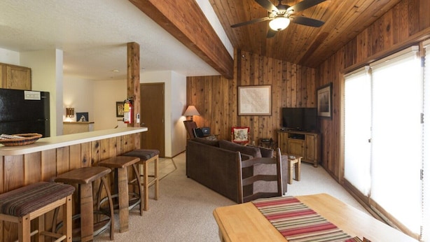 L’intérieur d’un chalet rustique est doté d’un plafond en bois, d’un coin bar avec des tabourets, d’un canapé et d’un téléviseur.