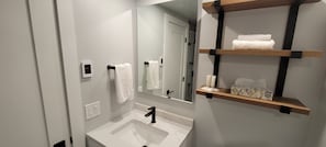 Et moderne badeværelse med en hvid vask, et spejl og en håndklædestang. Der er hylder med toiletartikler, og et brusebad er synligt i baggrunden.