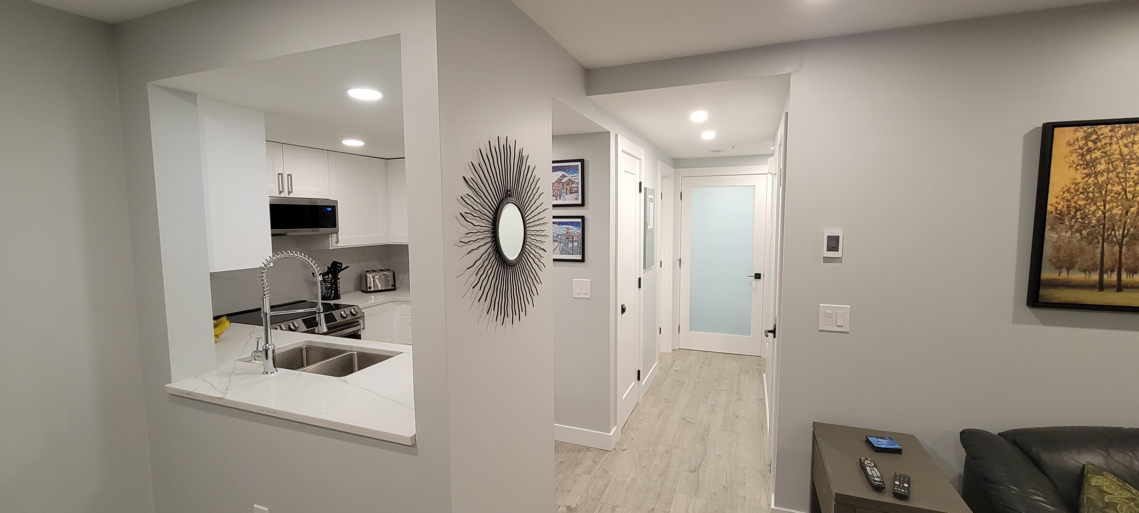 A modern kitchen with a built-in oven, a microwave, and a sink. There is a hallway leading to a bedroom with a door and a painting on the wall.
