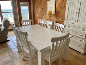 A dining area with a white table and chairs, a wooden wall, a lamp, and a view of the sea through a window. - Waterside: Soundfront condo with spectacular sunsets. (Ocracoke)