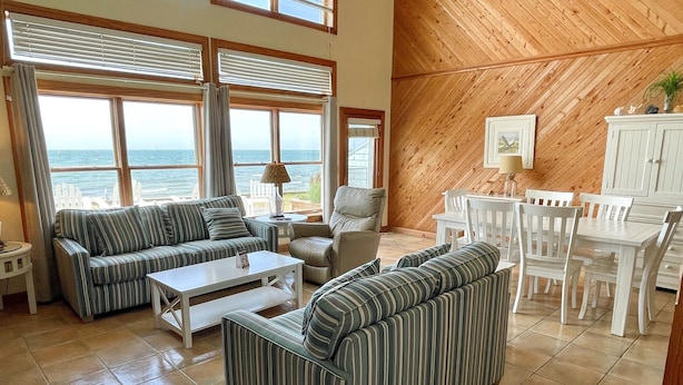 A spacious living room with a large window overlooking the sea, a dining area with white furniture, and a wooden ceiling. - Waterside: Soundfront condo with spectacular sunsets. (Ocracoke)