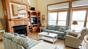 A living room with a fireplace, striped sofas, a TV, and a view of the sea through large windows. - Waterside: Soundfront condo with spectacular sunsets. (Ocracoke)