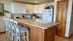 A kitchen with a wooden island, white bar stools, a microwave, a refrigerator, and a wooden door. - Waterside: Soundfront condo with spectacular sunsets. (Ocracoke)