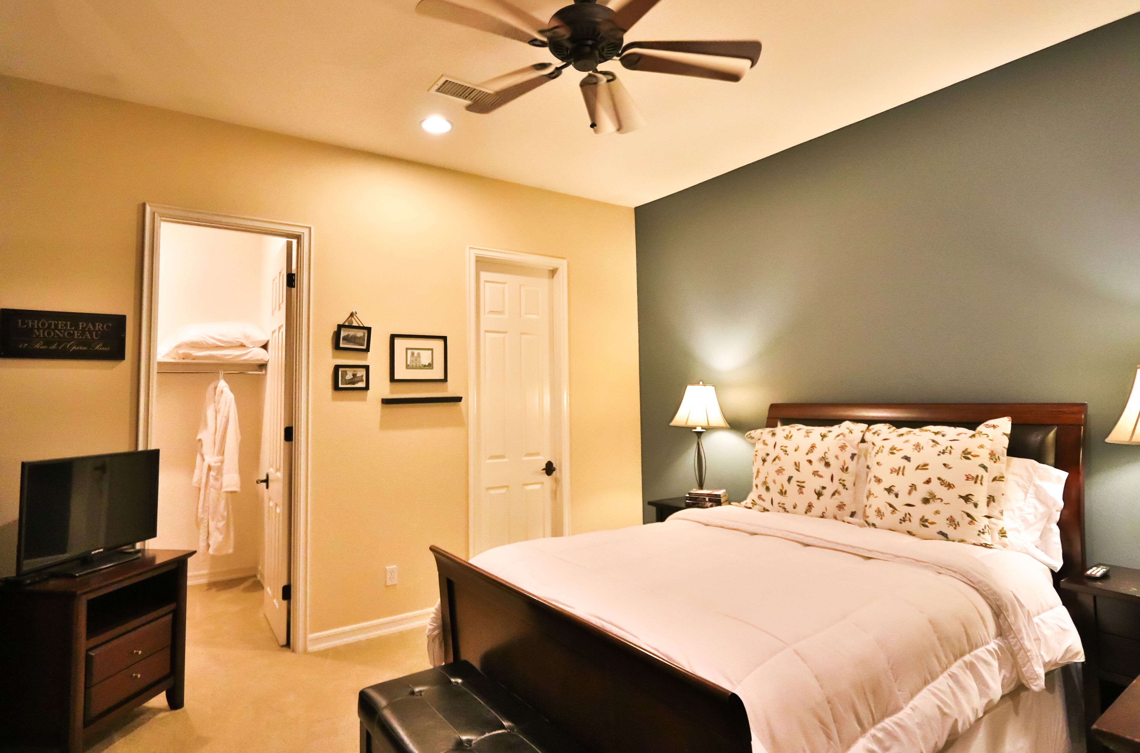 Habitación de hotel con cama, televisión, ventilador de techo, mesita de noche y puerta que da a otra habitación.