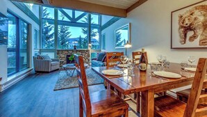 Uma sala de jantar com uma mesa de madeira posta para quatro pessoas, janelas amplas e uma lareira de pedra.