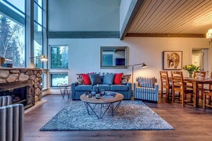 A spacious living area with a fireplace, a sofa with colorful cushions, a dining table, and a large window providing a view of the outdoors.