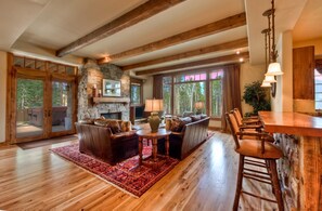 A spacious living room with a stone fireplace, wooden beams, large windows, and a bar area.