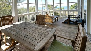 Een overdekt terras buiten met houten meubels, waaronder een tafel en stoelen, en uitzicht op de oceaan.