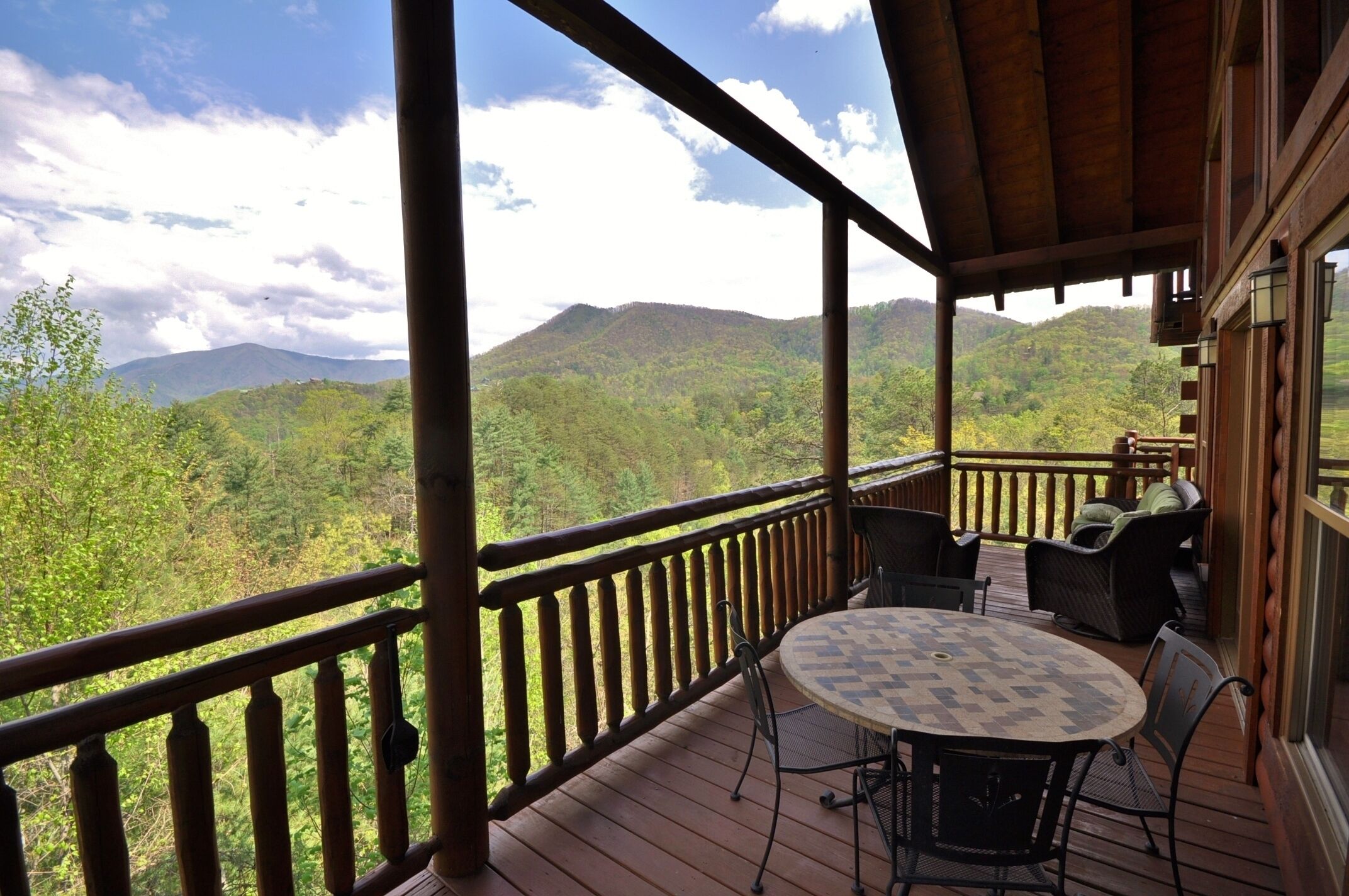 A wooden deck with a round table and chairs, overlooking a forested mountain range.