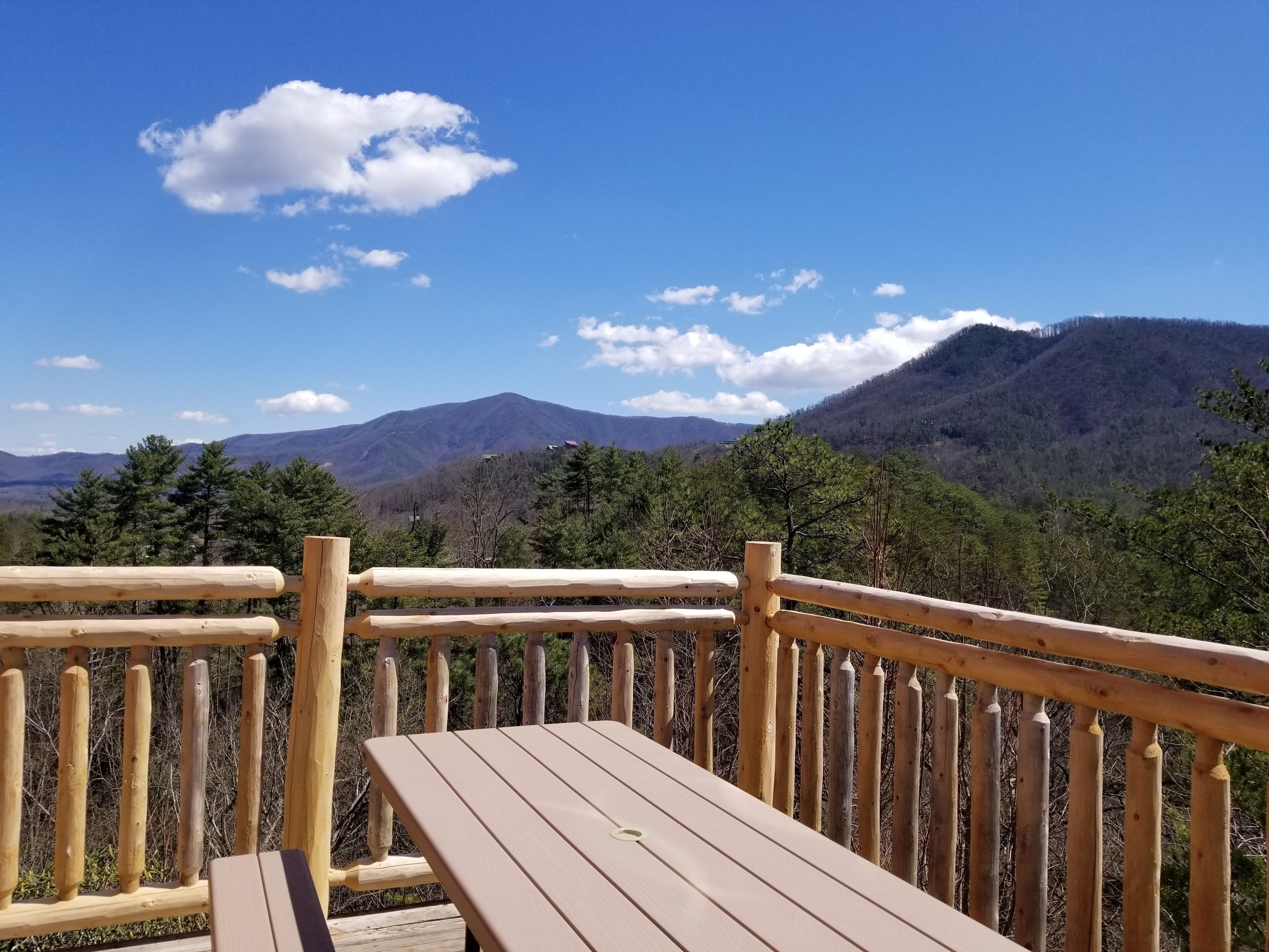 Eine Holzterrasse mit Tisch und Geländer, mit Blick auf eine Berglandschaft mit Bäumen und blauem Himmel.