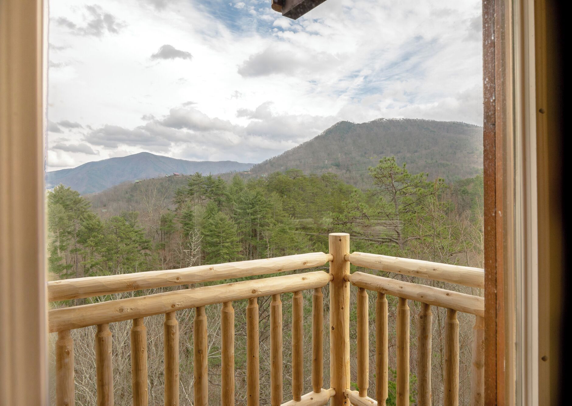 A wooden balcony railing overlooking a mountainous landscape with trees and a cloudy sky.