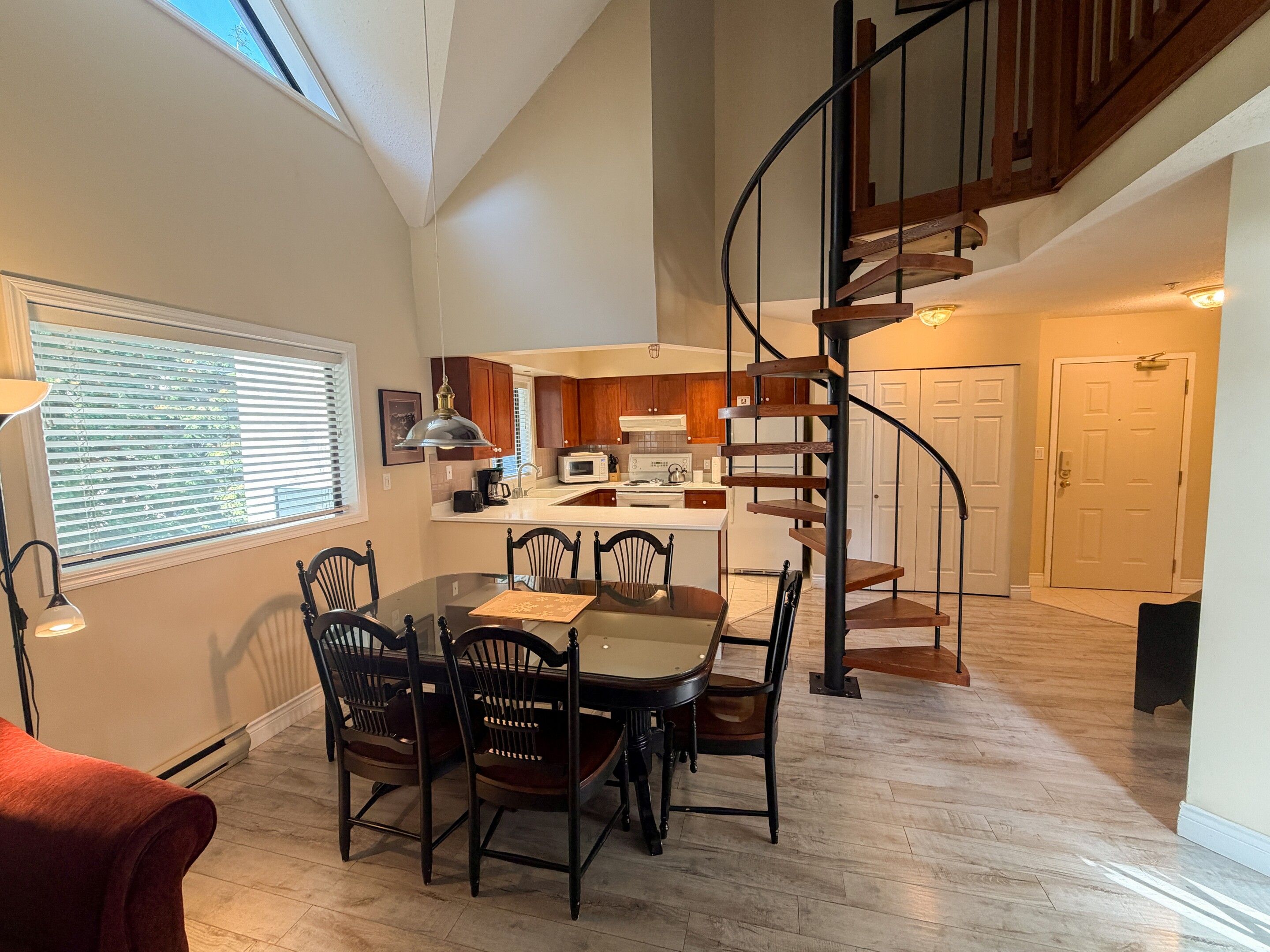 A dining area with a wooden table and chairs, a kitchen in the background, and a spiral staircase leading to an upper level.