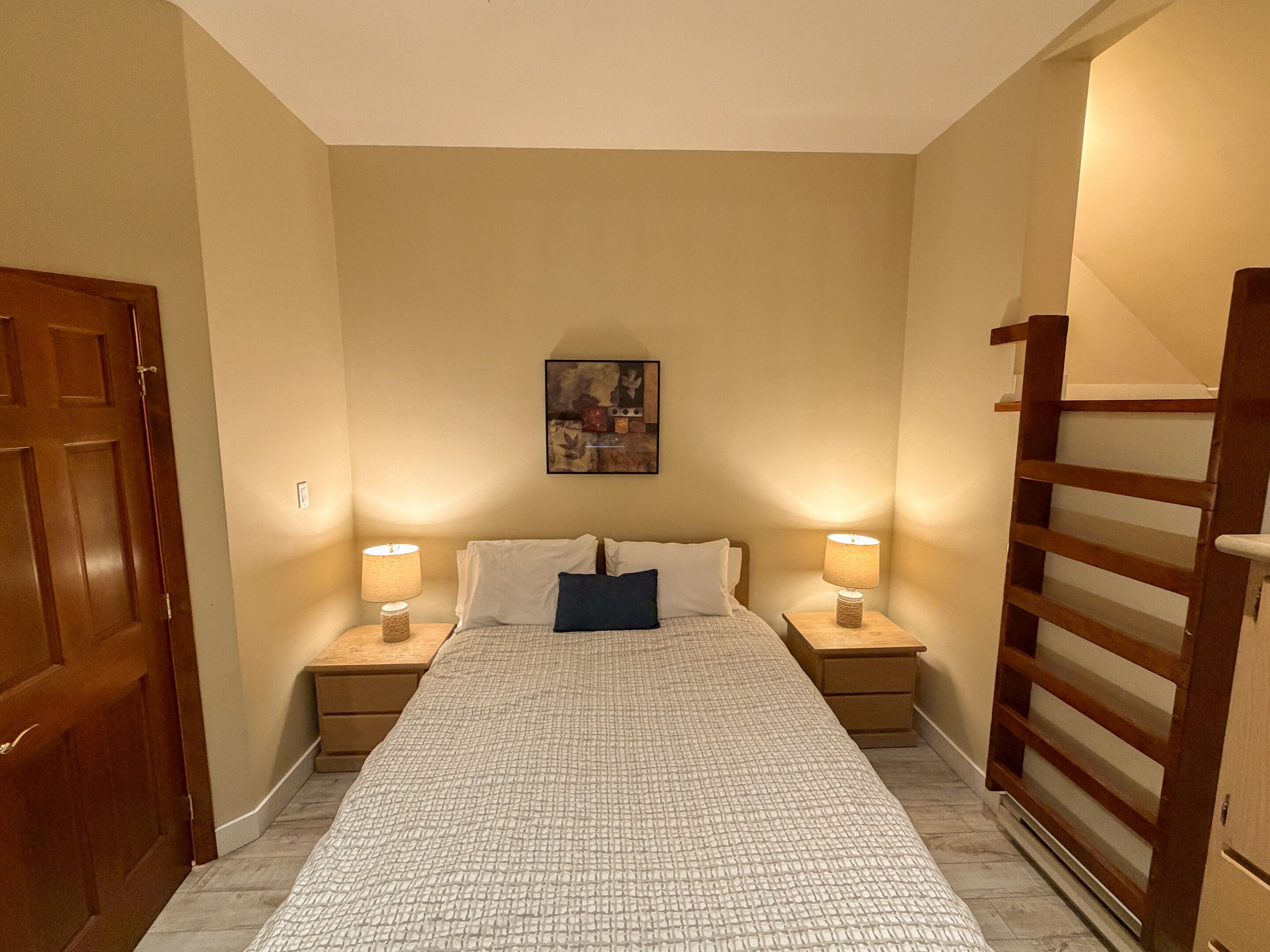 Una habitació d'hotel amb un llit, dues taules de nit, un quadre a la paret i una porta de fusta.