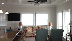 A spacious living area with a large wooden ceiling, a ceiling fan, and a television mounted on the wall. There are two windows with blinds, a sofa with floral cushions, and a dining area with a long table and chairs. - Shore Haven - 2 Bedrooms, 2 Bath, Sleeps 10 - Beach Front! (Crystal Beach)