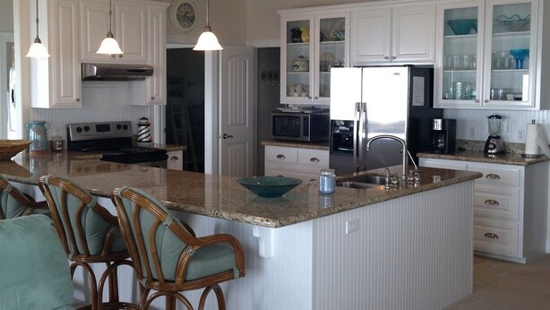 A spacious kitchen with white cabinetry, a central island with seating, and a dining area with wicker chairs. - Shore Haven - 2 Bedrooms, 2 Bath, Sleeps 10 - Beach Front! (Crystal Beach)
