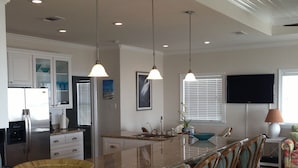 A spacious kitchen with a central island, bar stools, a refrigerator, and a television mounted on the wall. - Shore Haven - 2 Bedrooms, 2 Bath, Sleeps 10 - Beach Front! (Crystal Beach)