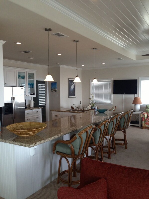 A spacious kitchen with a central island, bar stools, a refrigerator, and a television mounted on the wall. - Shore Haven - 2 Bedrooms, 2 Bath, Sleeps 10 - Beach Front! (Crystal Beach)