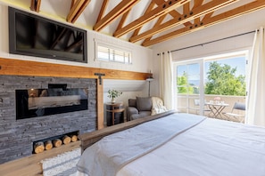 A bedroom with a stone fireplace, a large bed, a TV mounted on the wall, and a balcony with outdoor seating. - Ballard Canyon Ranch - Beautiful Home on 20 acres in Wine Country (Solvang)