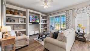 A living room with a beige sofa, a wooden coffee table, and a television on a stand. Shelves with decorative items and a window with curtains are also visible. - Liberty Sky: Private Pool, Water Park, Golf View, Near Disney (Kissimmee)