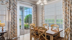 A dining area with a wooden table and chairs, large windows with blinds, and curtains with a floral pattern. - Liberty Sky: Private Pool, Water Park, Golf View, Near Disney (Kissimmee)