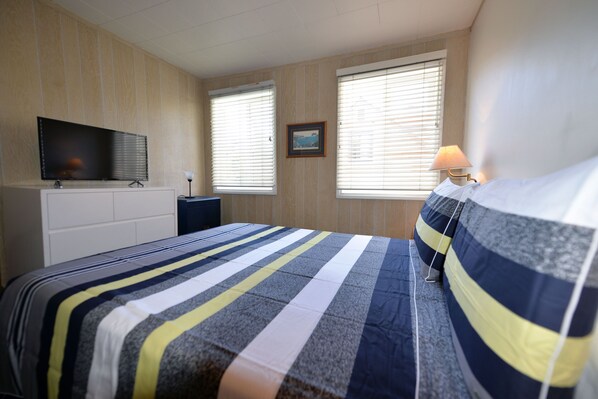 A bedroom with a bed, a TV, a lamp, and two windows with blinds.