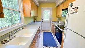 A kitchen with wooden cabinets, a white refrigerator, a built-in oven, and a window providing a view of trees. - Secret Cedars: Soundfront, perfect kayaking or water activities. (Ocracoke)