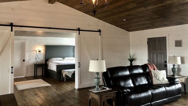 A spacious room with a wooden ceiling, a black leather sofa, a bed with white bedding, and a chandelier. - The PearTree Cottage- enjoy the view in the hot tub! 10 minutes from Berlin (Dundee)