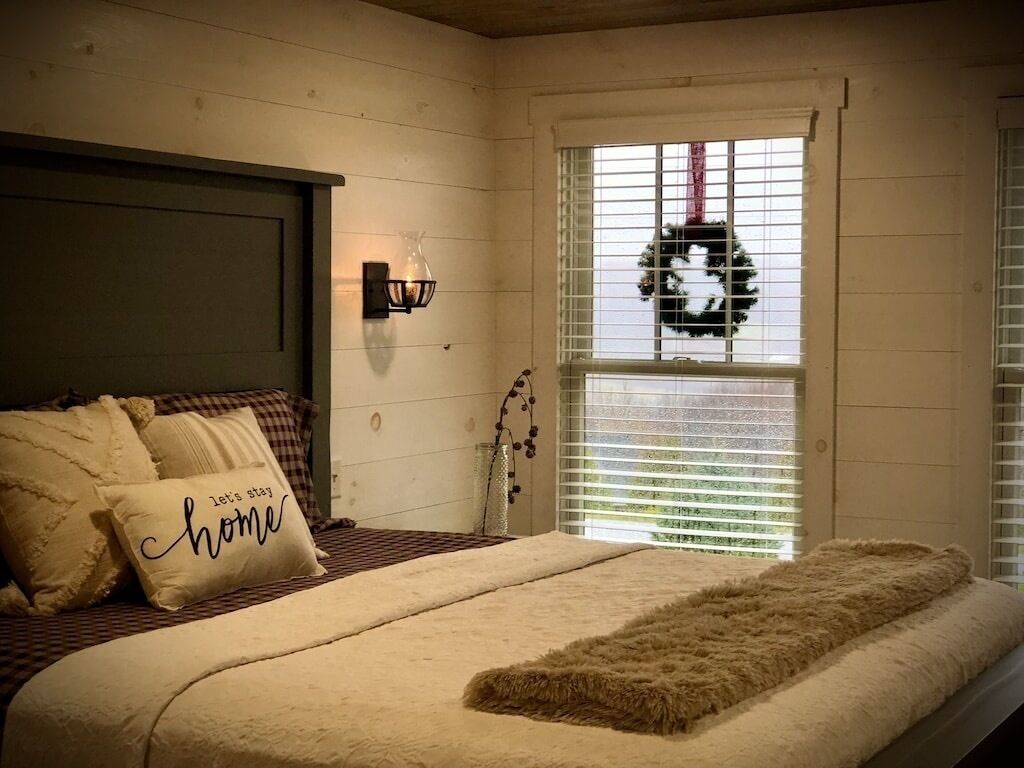 Quarto com cama, janela com persianas, luminária fixada na parede e um arranjo decorativo.