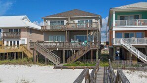 A beachfront property with a wooden deck leading to a house with multiple balconies and a covered outdoor area. - “Dolphin Watch” Amazing Gulf Front Home, Elevator, Paddleboard! (Navarre Beach)