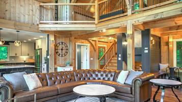 A spacious living area with a large leather sectional sofa, a round coffee table, and a dining table with chairs. A wooden staircase connects the upper and lower levels.