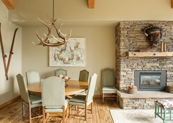 A dining area with a round wooden table, six upholstered chairs, a chandelier, a stone fireplace, and a mounted animal head. - Steps to Six Shooter Lift Access | Views (Big Sky)