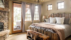 A rustic bedroom with a stone fireplace, a wooden bench, a bed with a large headboard, and a chandelier. - Steps to Six Shooter Lift Access | Views (Big Sky)