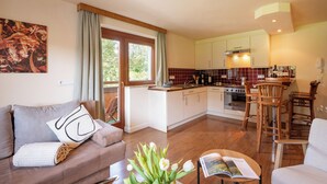 A modern living room with a sofa, a coffee table with a magazine, and a vase with flowers. There is a kitchen in the background with wooden cabinets and a dining area with chairs. - "Stylish and Comfortable One bedroomed Apartment in söll - Sleeps up to 4!" (Söll)