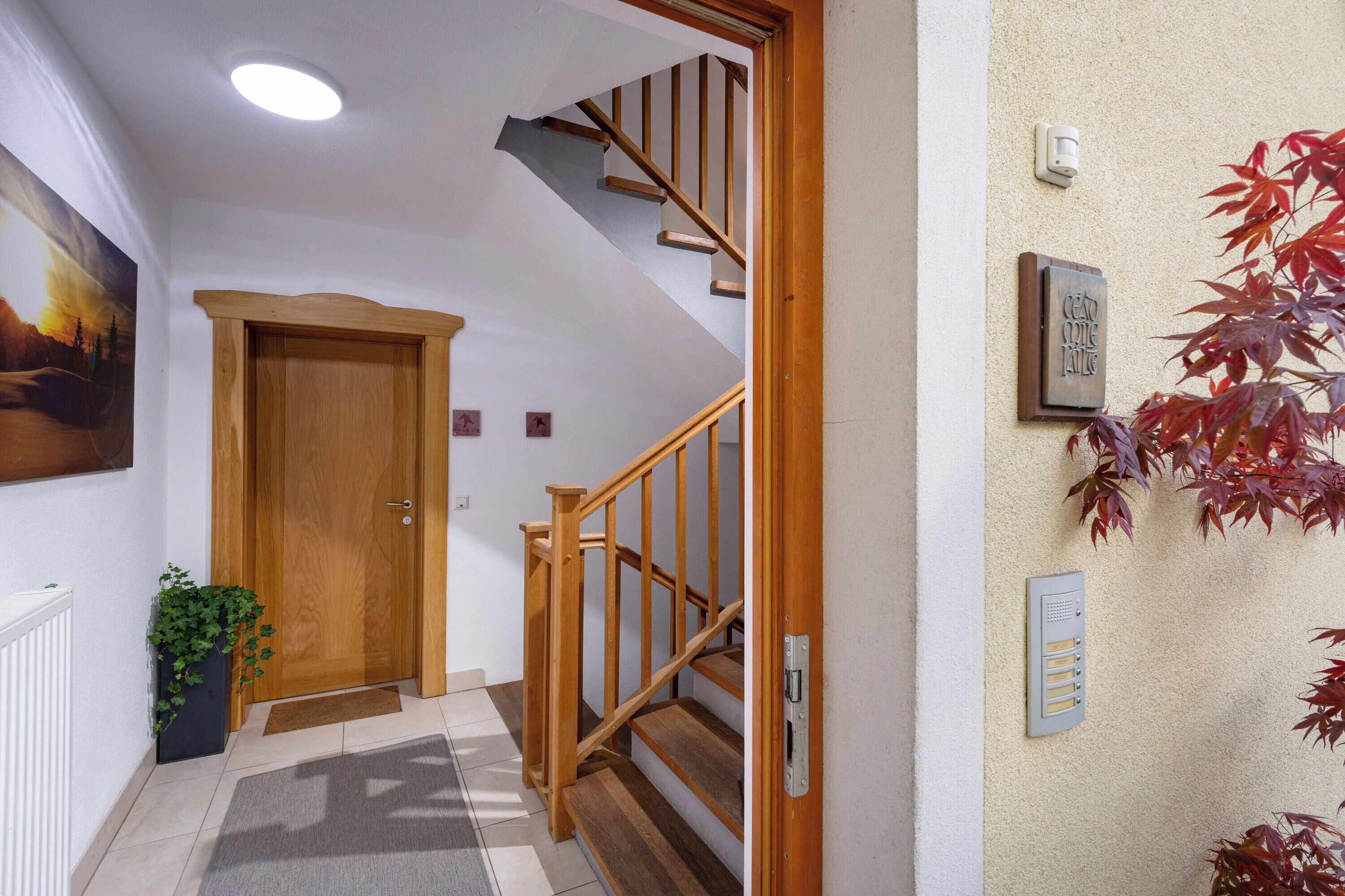 Una escalera de madera con barandilla, una puerta de madera, un portero automático en la pared y una planta en maceta.