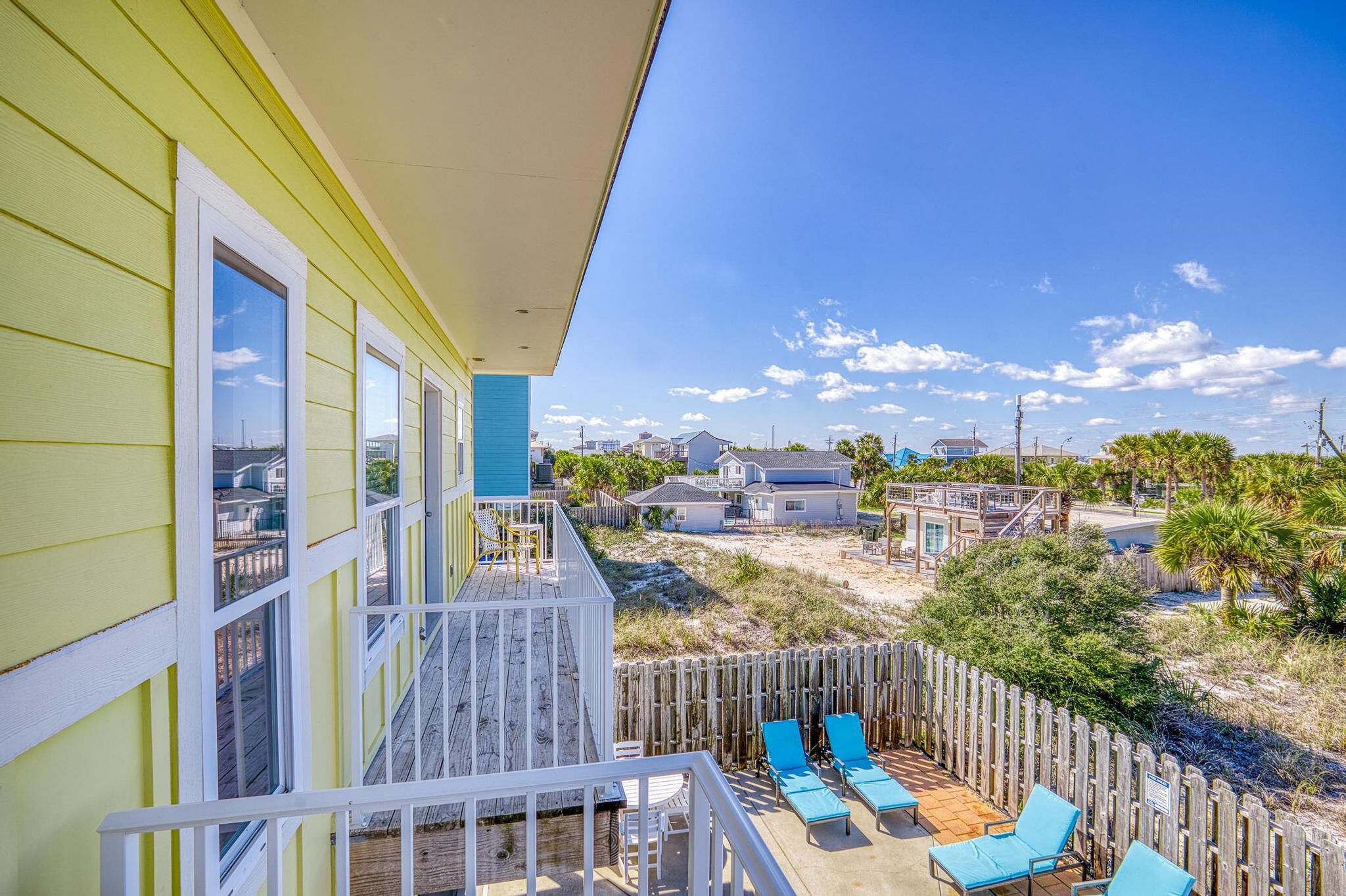 A balcony with a wooden deck, blue lounge chairs, and a view of a sandy beach and houses in the distance.