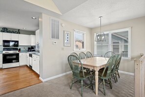 A compact kitchen with white cabinets, a microwave, and a stove. There is a dining area with a wooden table and green chairs. - Dolphin Watch: Oceanside, Village at Nags Head (Nags Head)