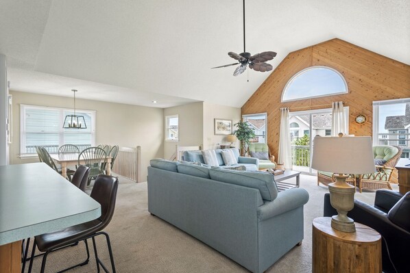 A spacious living area with a large sofa, dining table, and chairs, a ceiling fan, and a view of the outdoors through a large window. - Dolphin Watch: Oceanside, Village at Nags Head (Nags Head)