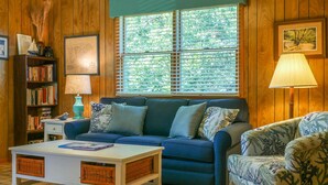 A cozy living room with a blue sofa, a white coffee table, wicker baskets, a bookshelf, and a window with blinds. - Lindley:  Near restaurants, spacious screened porch, rooftop deck. (Ocracoke)