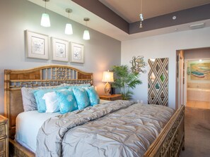 A bedroom with a wooden headboard, a bed with a grey quilt, and a nightstand with a lamp. There are framed pictures on the wall and a potted plant beside the bed.
