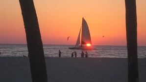 Atardecer sobre el océano con un velero y gente en la playa.