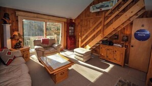Ein gemütliches Blockhaus-Interieur mit einer Couch, einem Sessel und einem kleinen Tisch. Eine Treppe mit Holzwand zu einem Balkon hinauf führt. Ein Schild mit der Aufschrift 'EVACUATION ROUTE' ist an der Wand sichtbar.