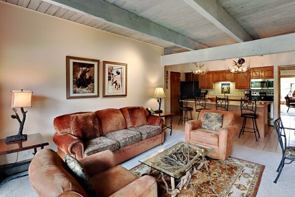 A spacious living room with a large sofa, a smaller armchair, and a coffee table. There is a kitchen in the background with wooden cabinets and a chandelier.