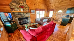 A cozy cabin interior with a stone fireplace, wooden floors, a red sofa, a television, and a kitchen area. - Absolute High | 3BR Cabin w/ Long-Range Mountain Views, Game Room & Deck (West Jefferson)