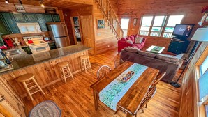 A cozy cabin interior with a wooden dining table, bar stools, a sofa, and a TV in the living area. - Absolute High | 3BR Cabin w/ Long-Range Mountain Views, Game Room & Deck (West Jefferson)