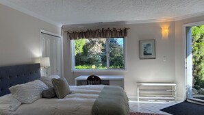 Ein Schlafzimmer mit Bett, Nachttisch, Lampe, Fenster und Blick auf Grünfläche.