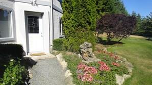 Ein Haus mit weißer Fassade, einer Haustür, einem kleinen Garten mit Blumen und Steinen sowie einem Baum mit violetten Blättern.